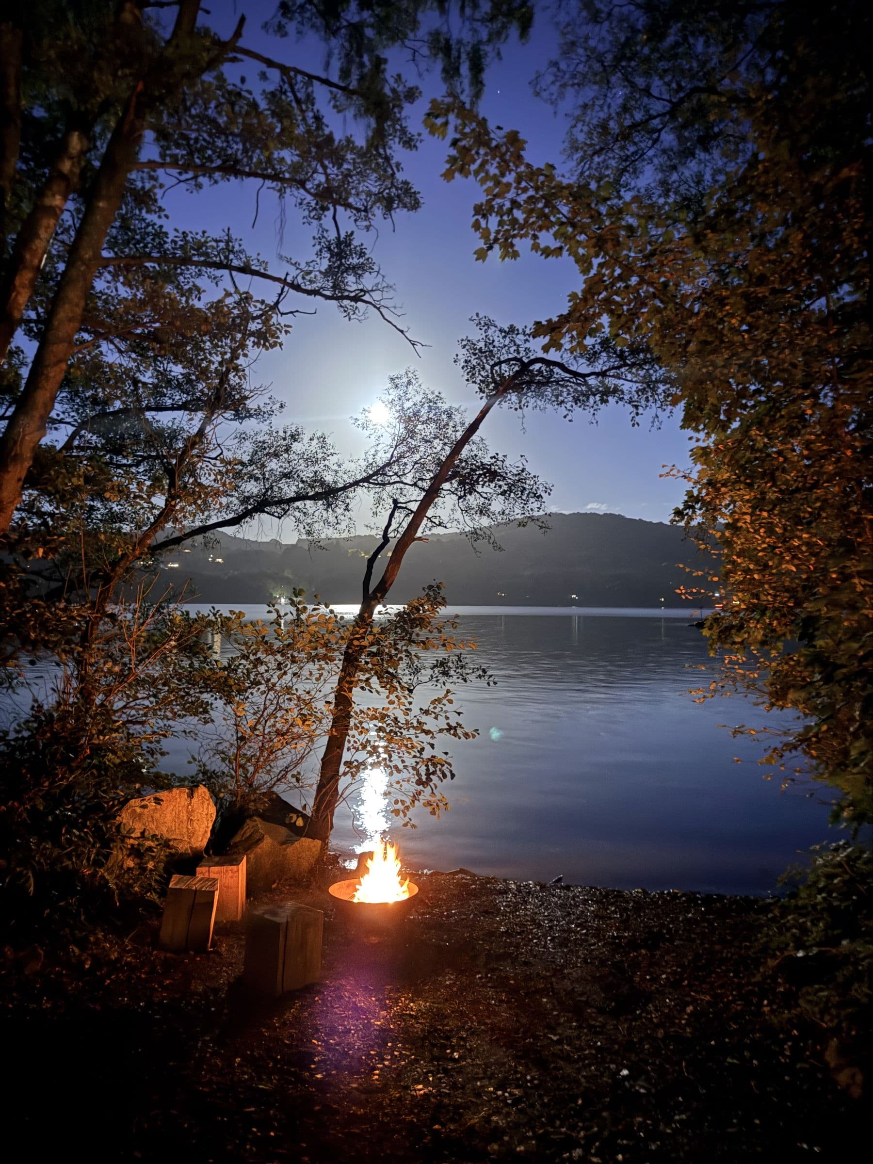 A firepit glowing on the shore at night beside wooden block seats, the moon over Windermere through the trees