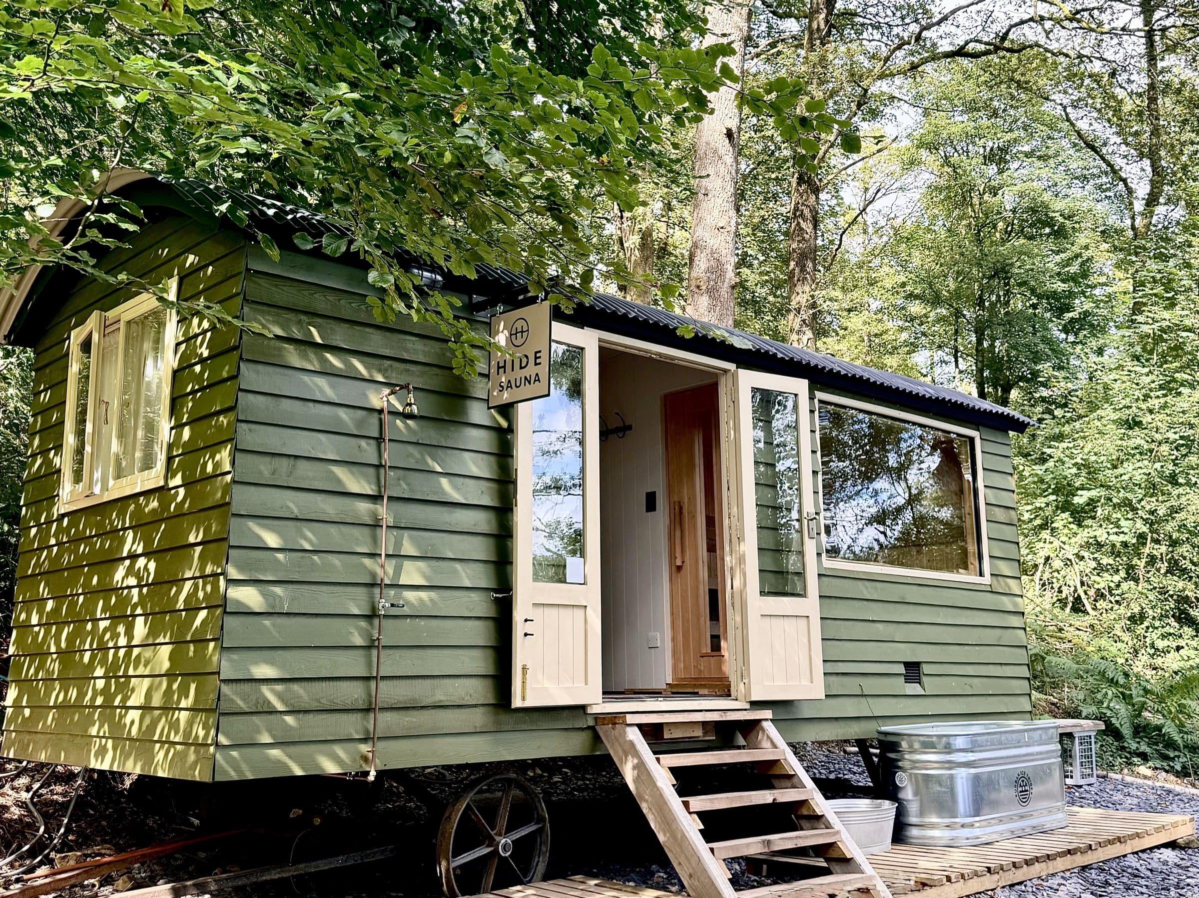 Hide Sauna's green timber cabin in dappled sunlight under the trees, doors open beneath the HIDE SAUNA sign, steps down to the plunge tub