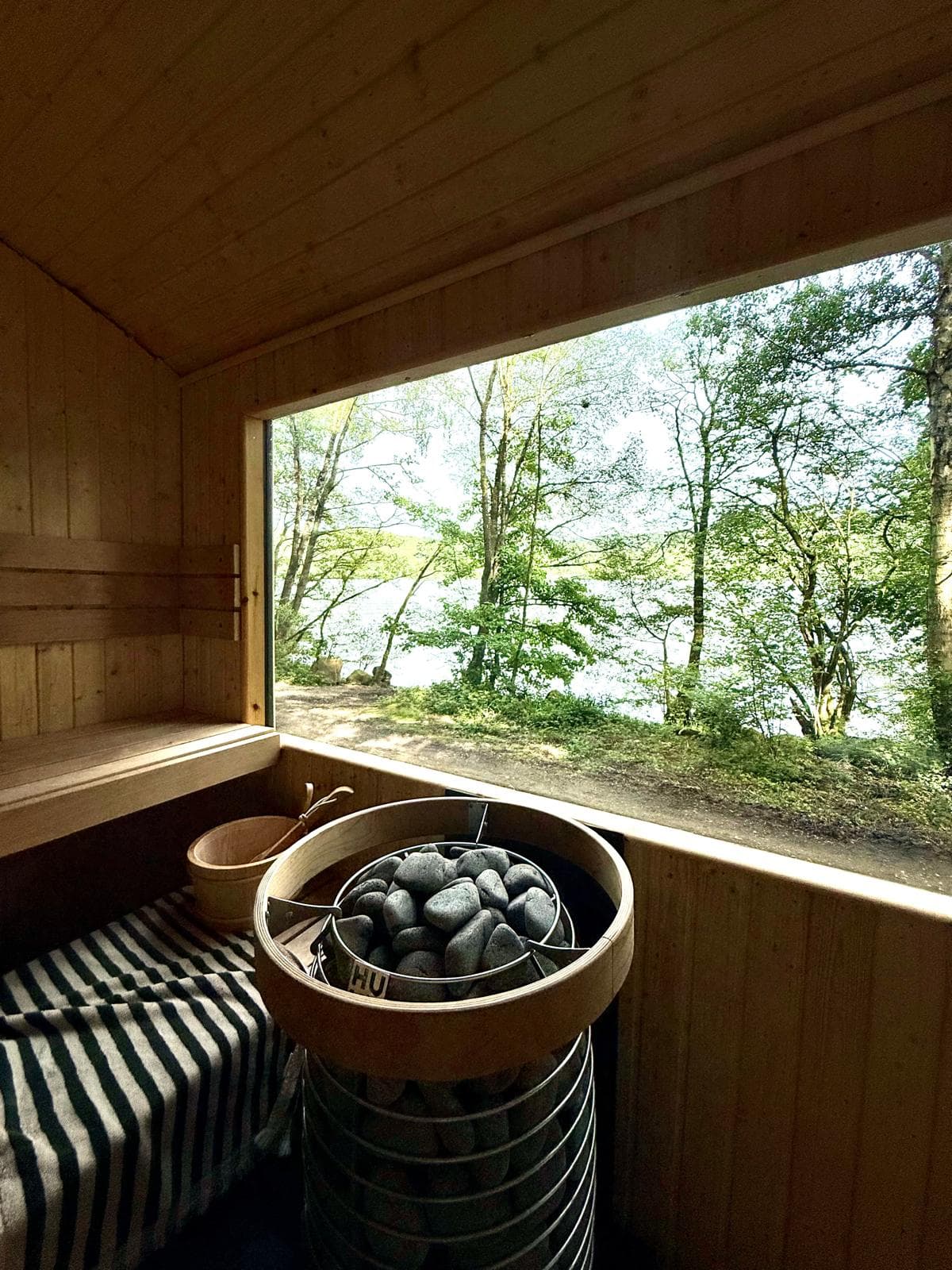 Inside the timber sauna: the stone heater beside the pine bench, the panoramic window looking out through trees to the lake